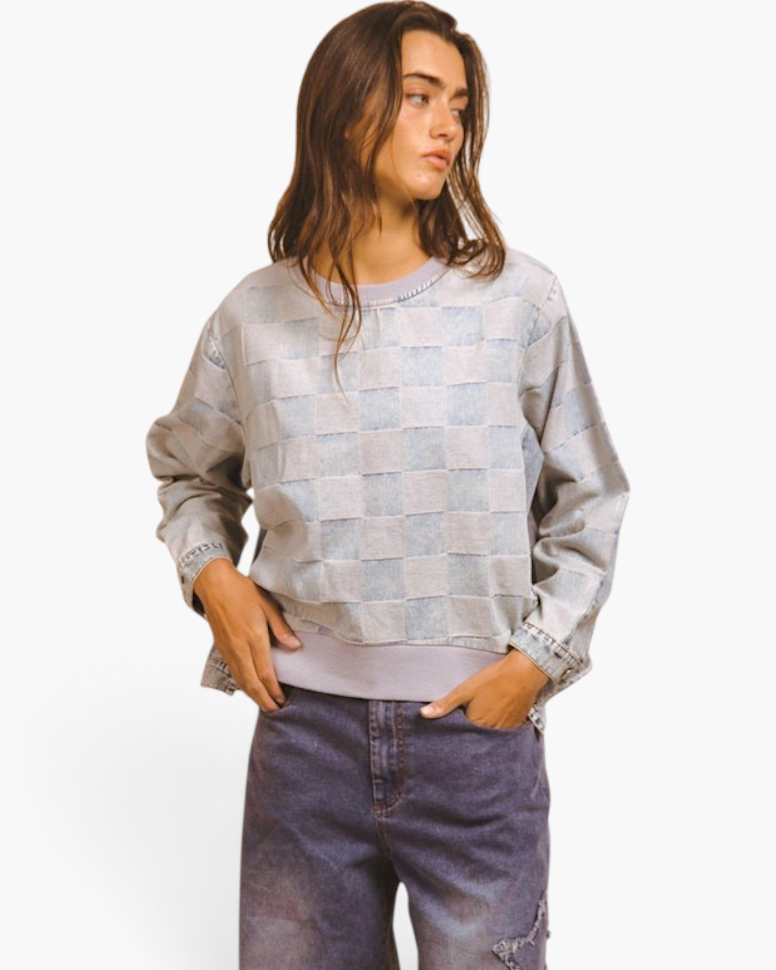 A woman wearing a light gray and blue checkerboard patterned sweatshirt with rolled cuffs, paired with dark blue jeans, against a plain white background.
