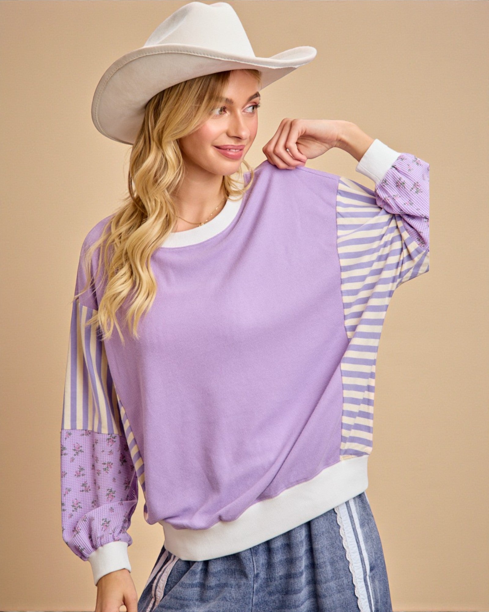 Woman wearing a purple sweatshirt with striped sleeves and a white cowboy hat on a beige background