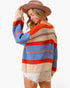 A woman wearing a loose knit sweater with horizontal stripes in beige, light blue, and orange-red, paired with white frayed shorts and a brown wide-brim hat, against a plain white background.