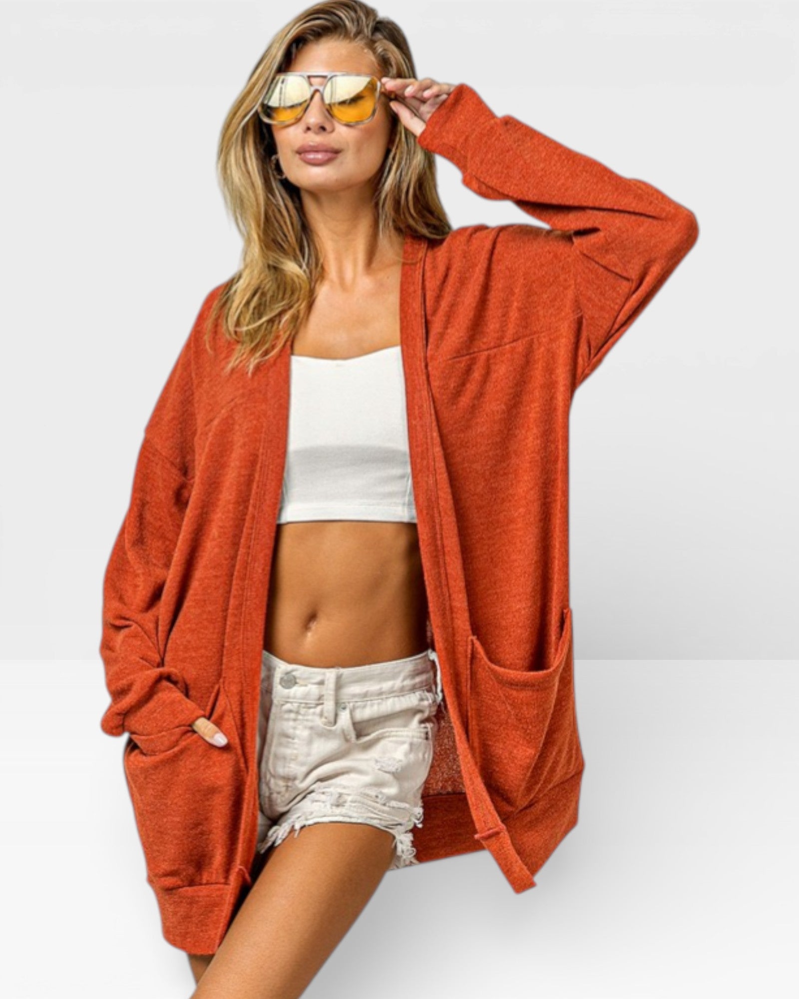 Woman wearing a rust-colored cardigan over a white top and shorts, with yellow sunglasses on a light gray background.