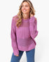 A woman wearing a chunky knit openwork sweater in a dusty purple color, paired with blue jeans, against a plain white background.