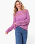 A woman wearing a lavender open-knit long-sleeve sweater and blue jeans, standing against a plain white studio background.