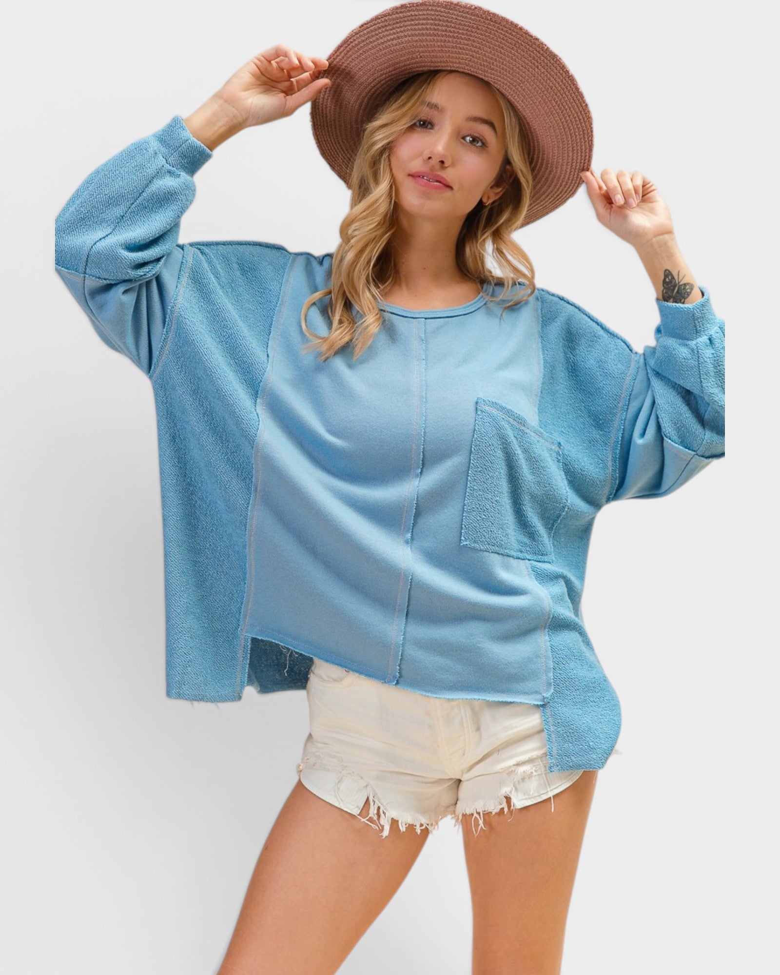 Woman wearing a blue oversized sweatshirt and white shorts with a brown hat on a plain background