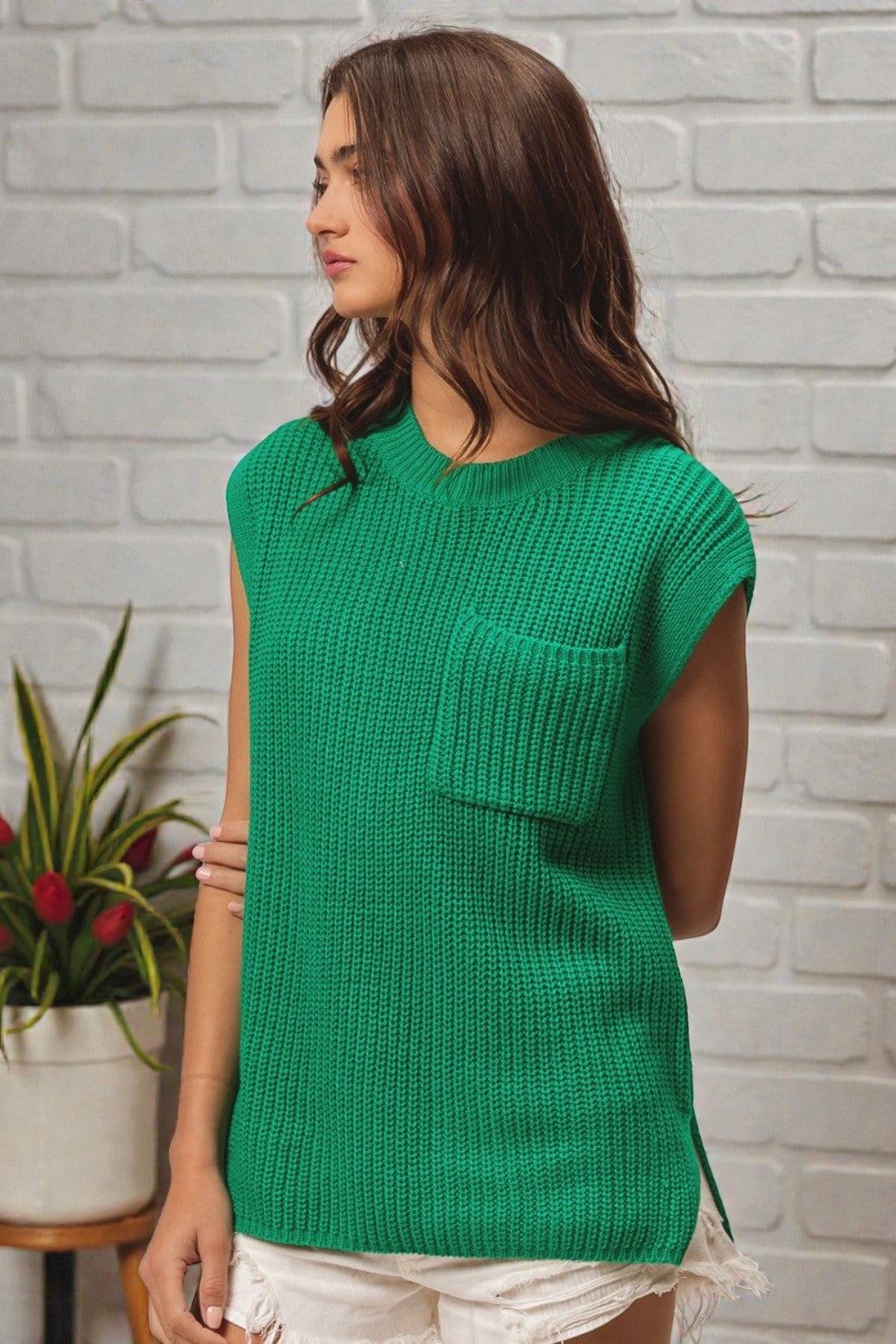 Woman wearing a green sleeveless knitted top against a white brick wall.