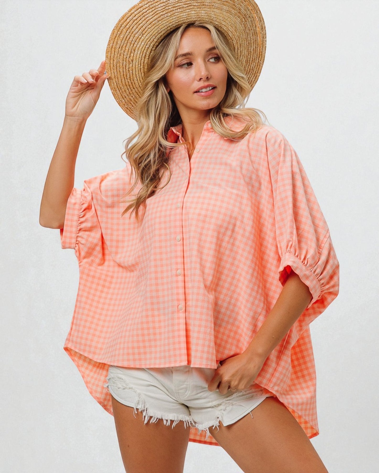 Woman wearing a peach-colored checkered cover-up, white shorts, and a straw hat on a plain background