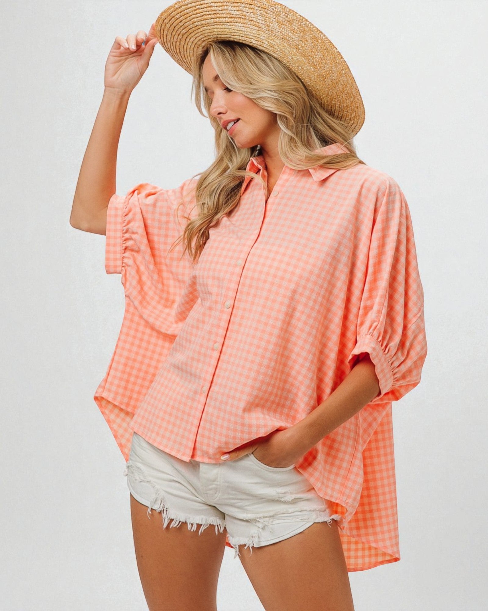 Woman wearing a peach-colored checkered shirt and white shorts, holding a straw hat against a plain background.