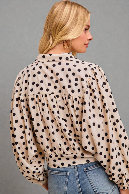 Woman wearing a beige blouse with black polka dots and blue jeans against a gray background