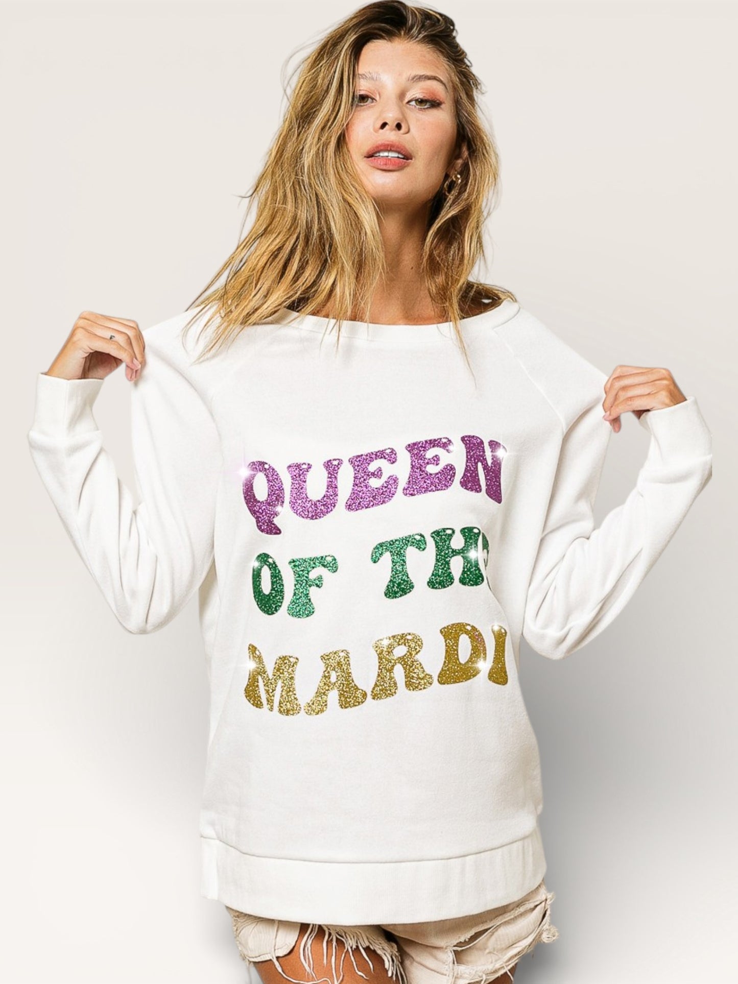 Woman wearing a white sweatshirt with colorful text 'QUEEN OF THE MARDI' on a plain background