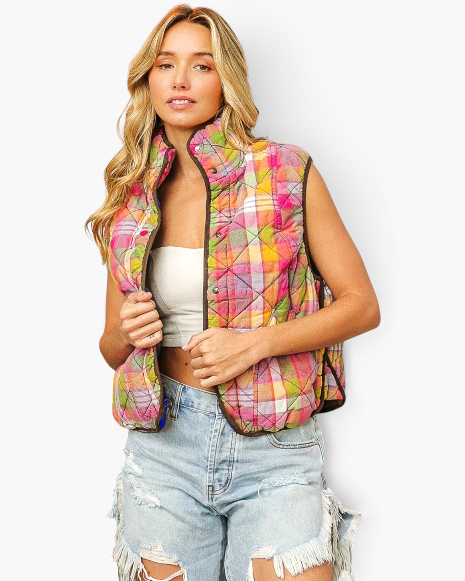 A woman wearing a sleeveless quilted puffer vest with a vibrant multicolor plaid pattern in pink, yellow, green, and orange, styled with a white crop top and distressed denim shorts against a light gray background.