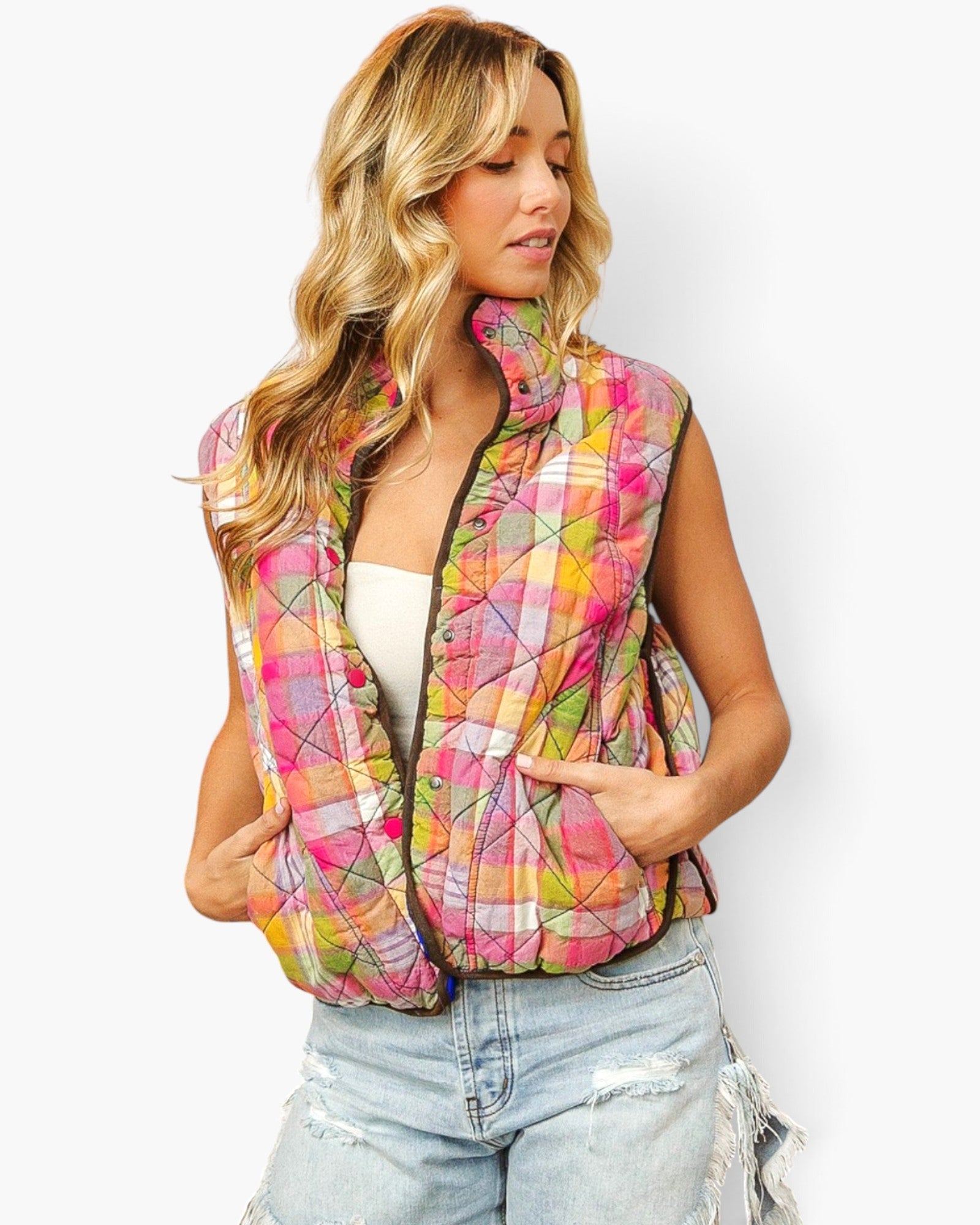 A woman wearing a sleeveless quilted puffer vest with a vibrant pink, orange, yellow, and green abstract plaid pattern, styled with light-wash distressed jeans against a plain white background.