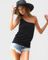 Woman wearing a black one-shoulder top and denim shorts with a wide-brimmed hat on a light gray background