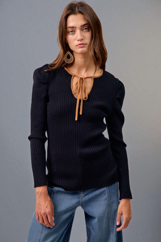 Woman wearing a black ribbed sweater with a brown tie and blue jeans against a gray background