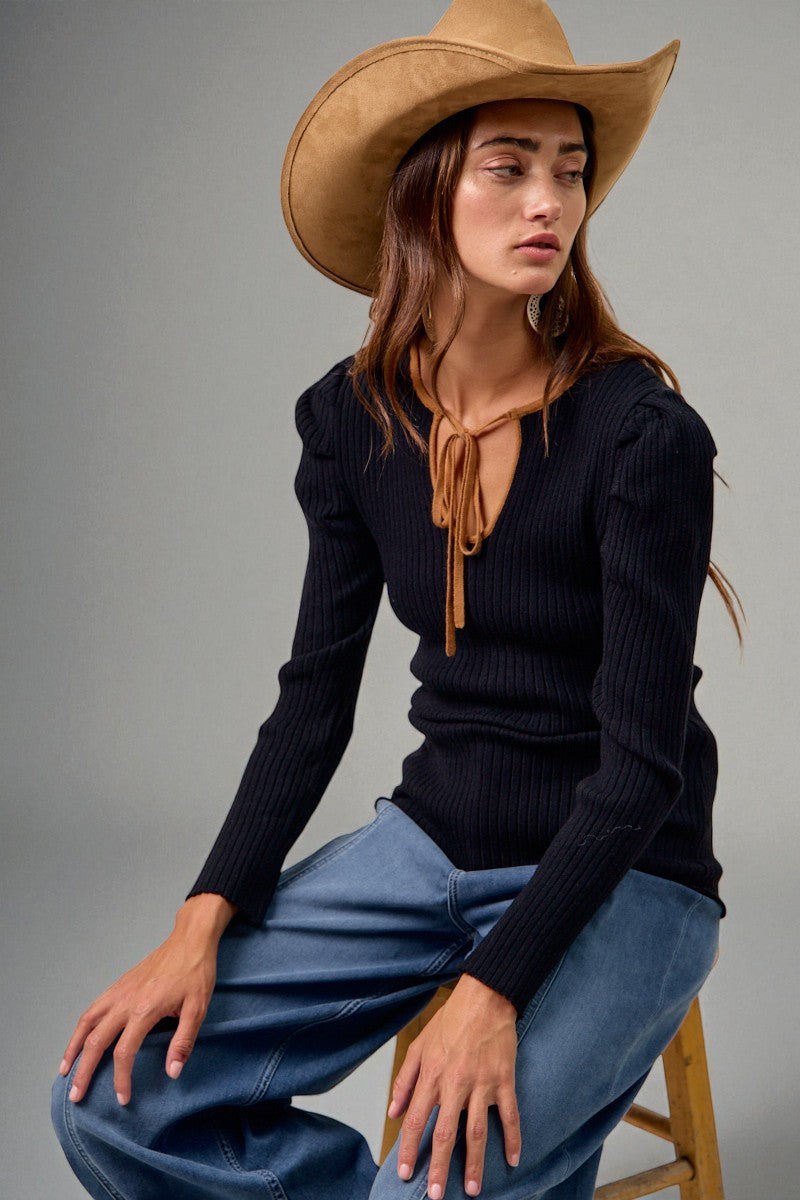 Woman wearing a black ribbed sweater, blue jeans, and a tan cowboy hat against a gray background