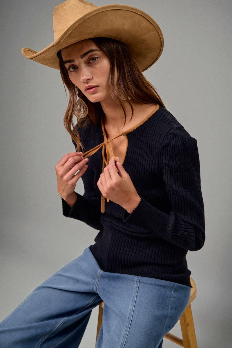 Woman wearing a black sweater, blue jeans, and a tan cowboy hat against a gray background