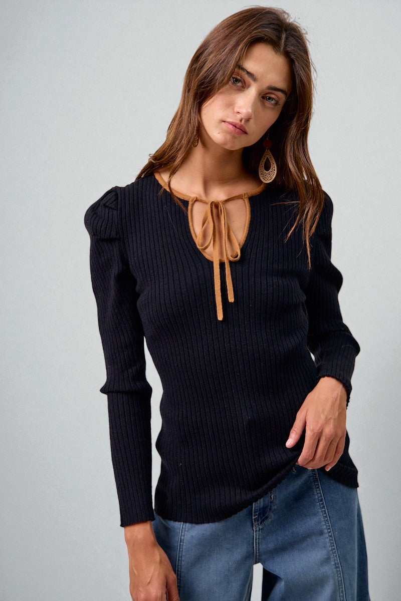 Woman wearing a black ribbed sweater with a tie detail and blue jeans on a light gray background