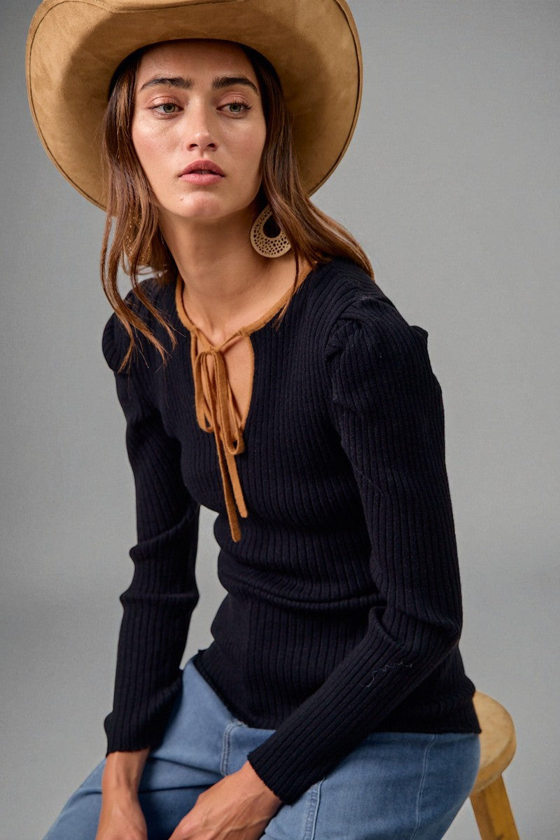 Woman wearing a black ribbed sweater, blue jeans, and a beige hat with a brown ribbon against a gray background.