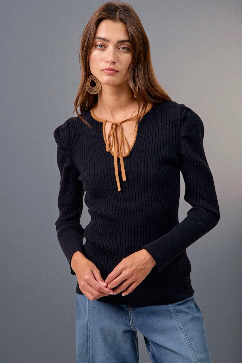 Woman wearing a black ribbed sweater with a tie detail and blue jeans against a gray background