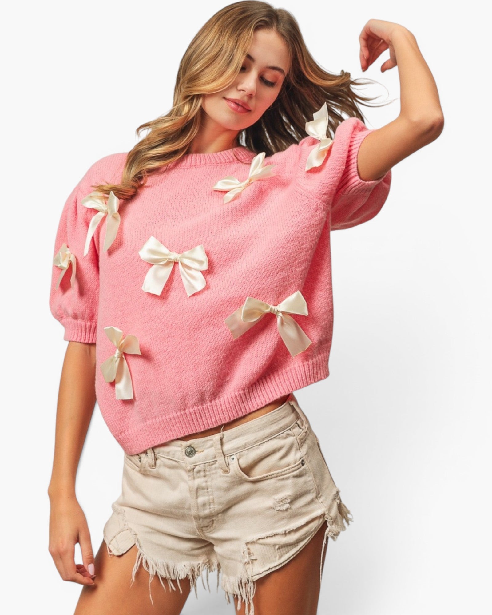 A woman wearing a pink short-sleeve knit sweater adorned with multiple cream satin bow appliqués, paired with light beige distressed denim shorts, against a plain white studio background.