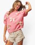 A woman wearing a pink short-sleeve knit sweater adorned with multiple cream satin bow appliqués, paired with light beige distressed denim shorts, against a plain white studio background.