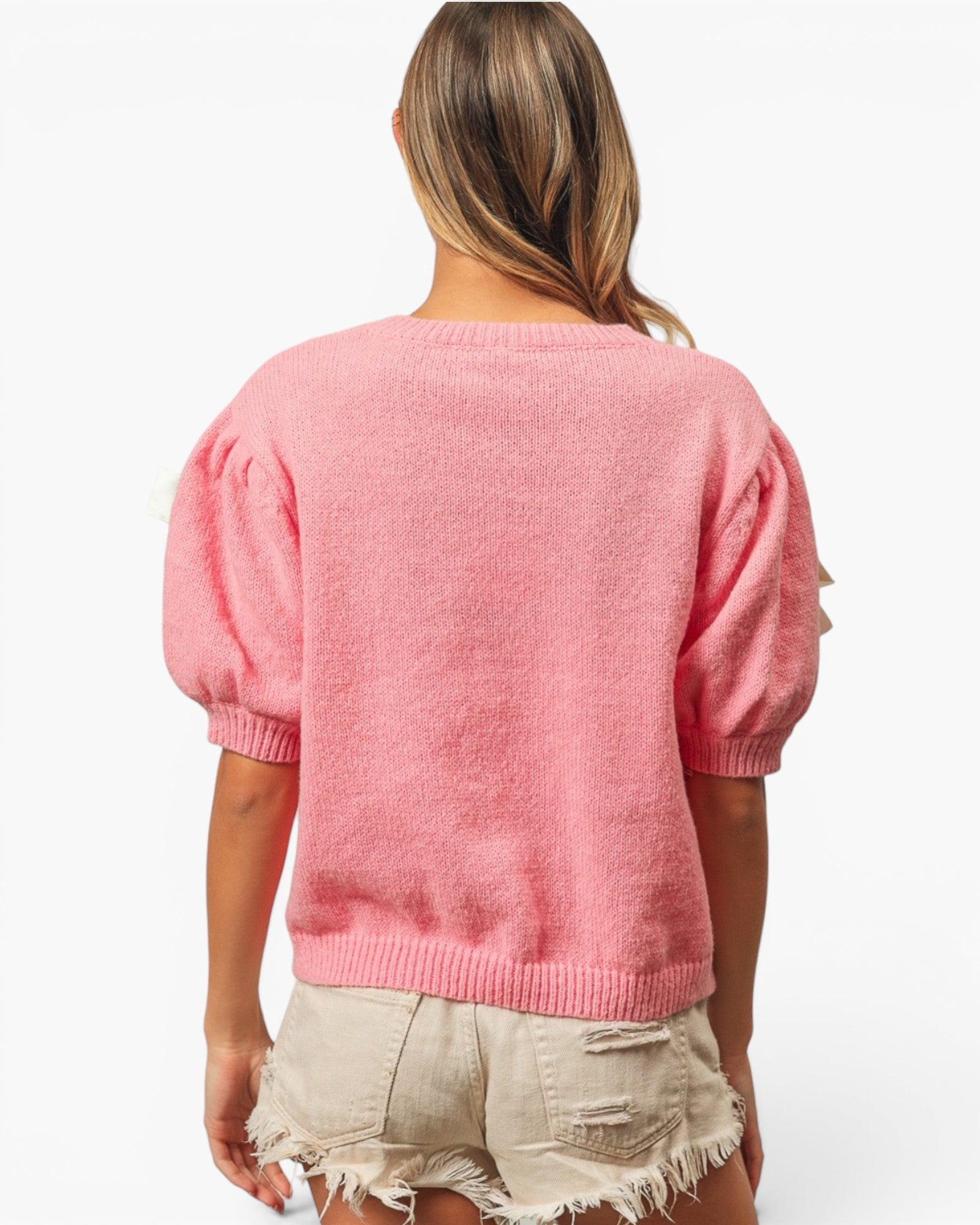 A woman shown from the back wearing a pink short-sleeve knit sweater with voluminous puff sleeves and beige frayed denim shorts against a plain white background.