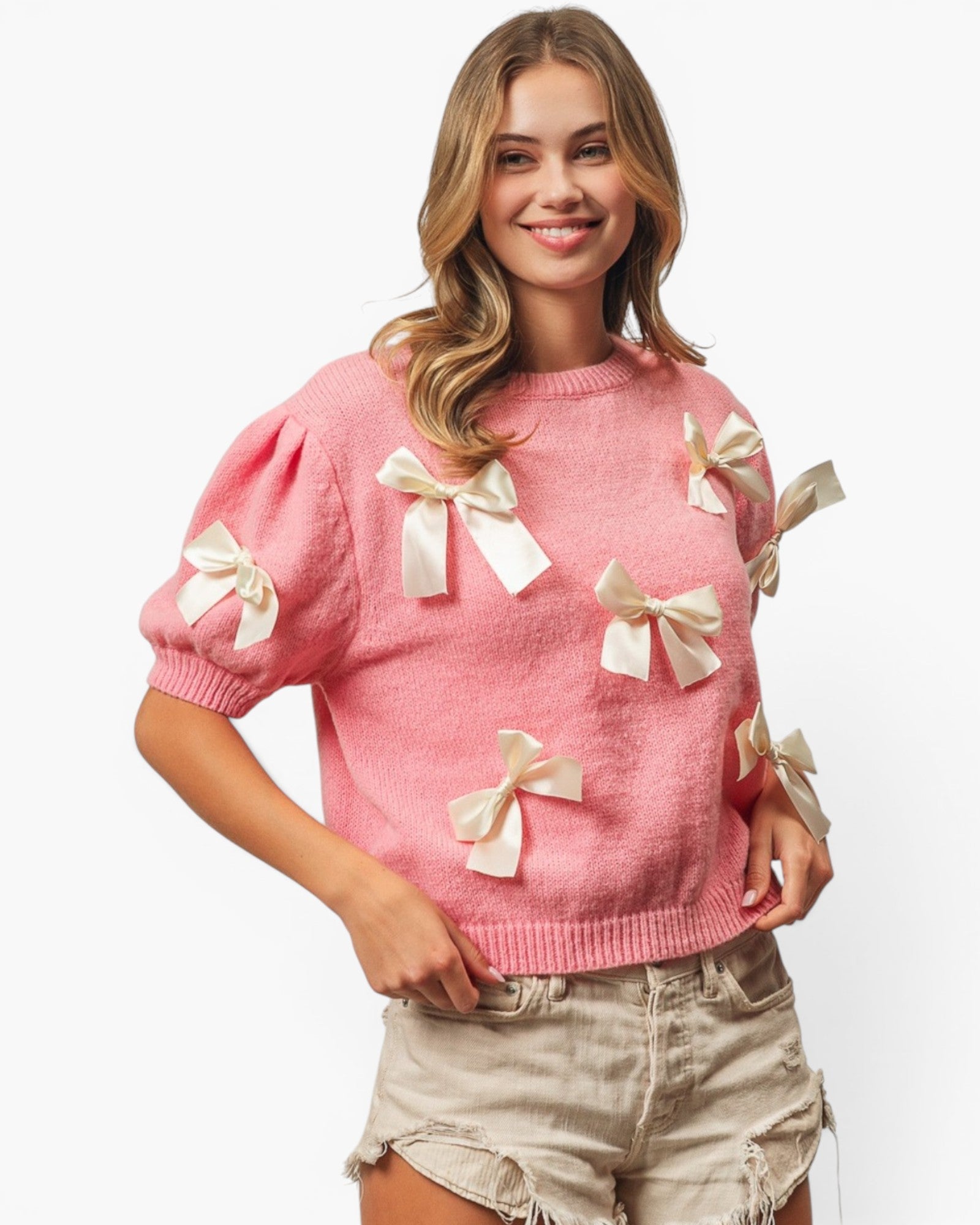 A woman wearing a pink knit sweater with cream satin bow details on the sleeves and front, paired with light beige denim shorts, against a plain white background.