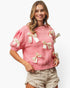 A woman wearing a pink knit sweater with cream satin bow details on the sleeves and front, paired with light beige denim shorts, against a plain white background.