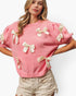 A woman wearing a pink knit sweater with cream satin bow details on the sleeves and front, paired with beige shorts, against a plain white background.