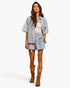 A woman modeling a matching two-piece outfit featuring a light blue and white floral print short-sleeve button-up shirt and shorts set, paired with a cream ribbed tank top and brown western boots, against a plain white studio background.