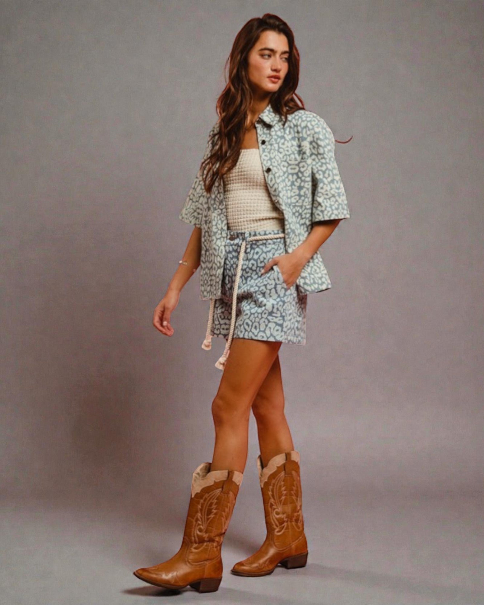 Woman wearing a patterned outfit with a white top, shorts, and brown cowboy boots on a gray background