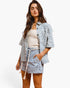 A woman modeling a matching light blue and white patterned short-sleeve button-up shirt and shorts set with a cream knit top, against a plain white studio background.