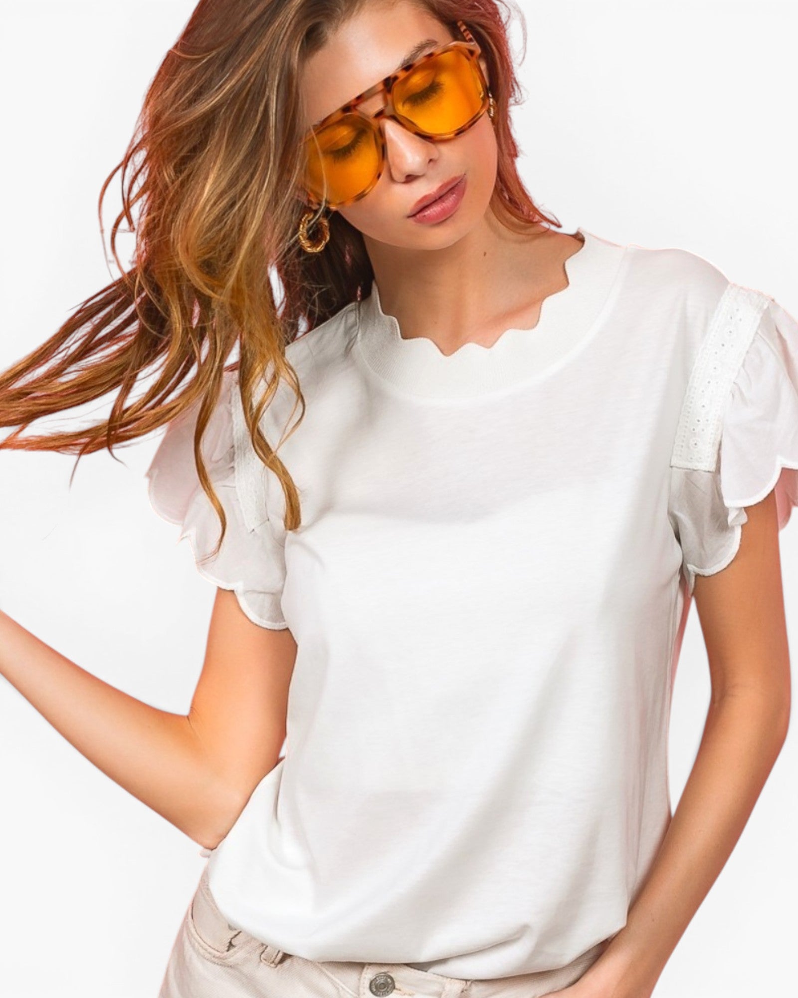 A woman wearing a white short-sleeve top with ruffled sleeves and a scalloped neckline, paired with beige pants and orange-tinted sunglasses, against a plain white background.