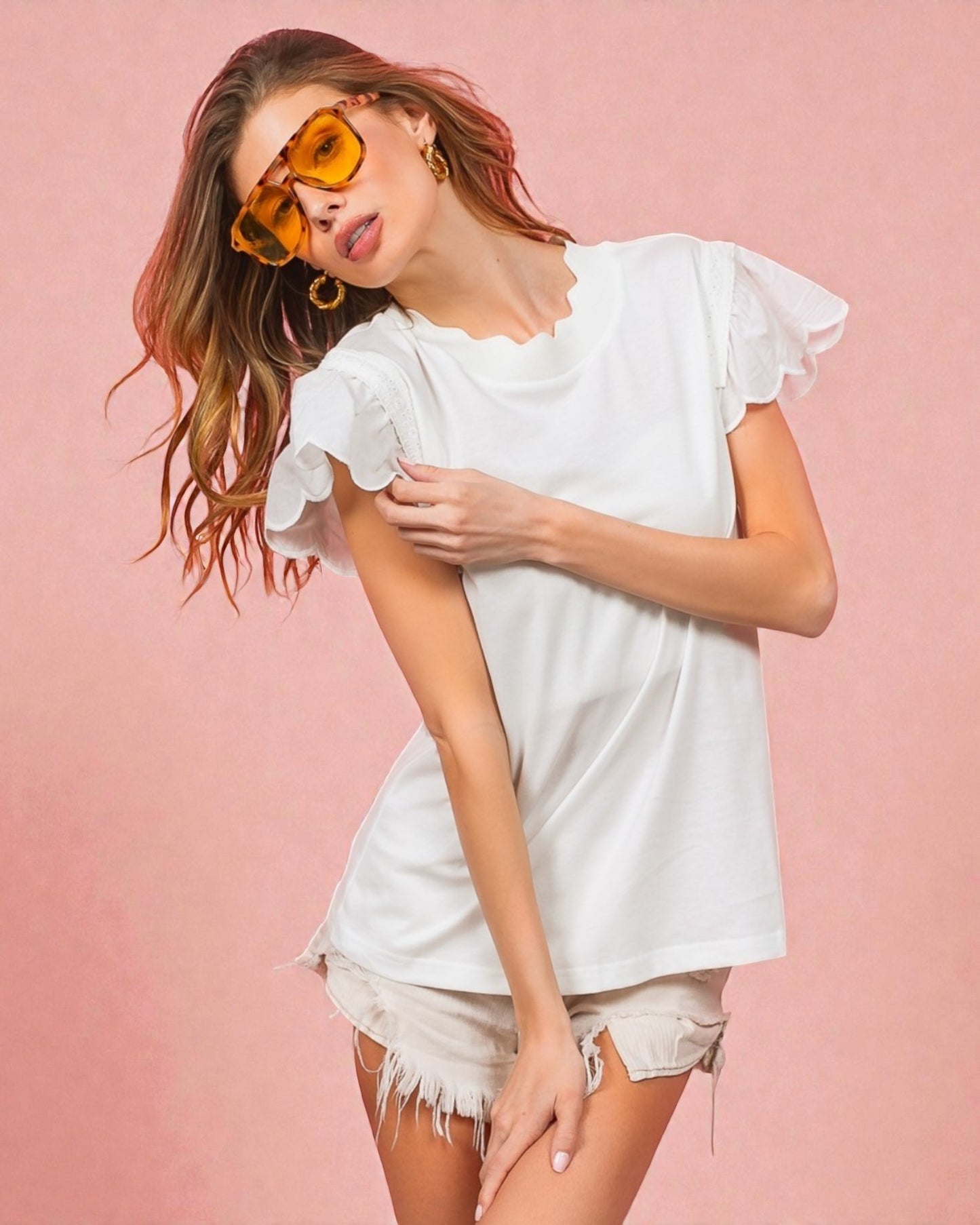 Woman wearing a white ruffled top and shorts with yellow sunglasses on a pink background