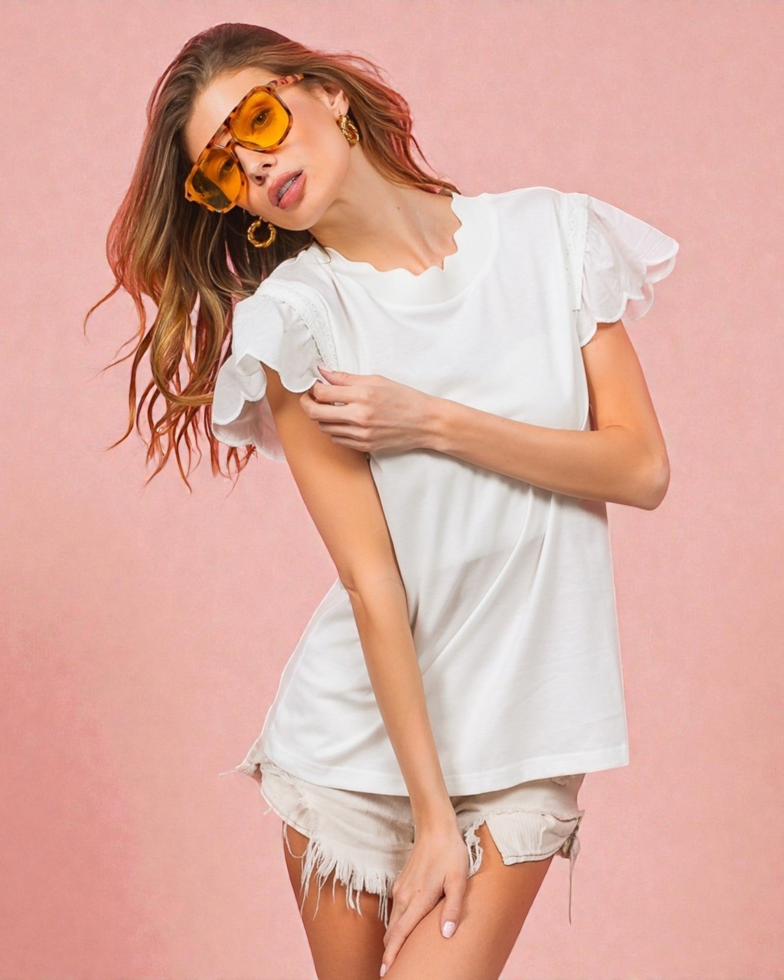 Woman wearing a white ruffled top and shorts with yellow sunglasses on a pink background
