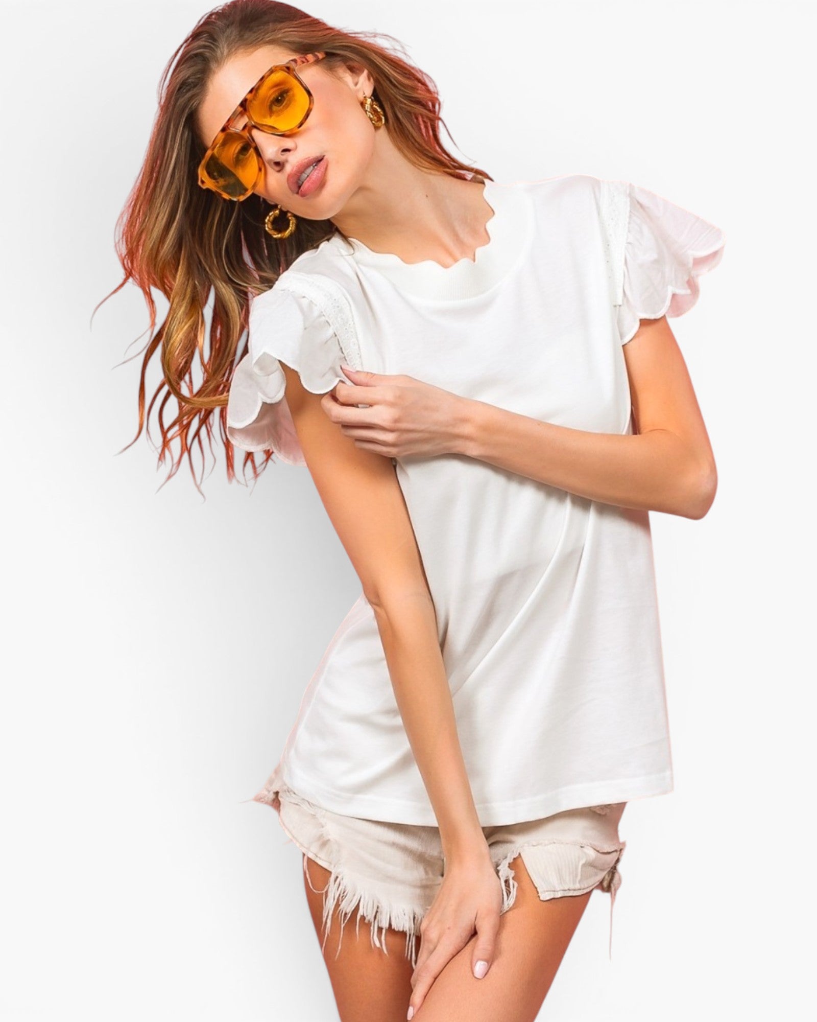 A woman modeling a white short-sleeve t-shirt with ruffled flutter sleeves and scalloped edges, paired with distressed denim shorts and yellow-tinted sunglasses, against a plain white background.