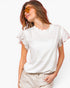 A woman wearing a white short-sleeve blouse with ruffled flutter sleeves and a scalloped neckline, paired with light beige shorts, against a plain white background.