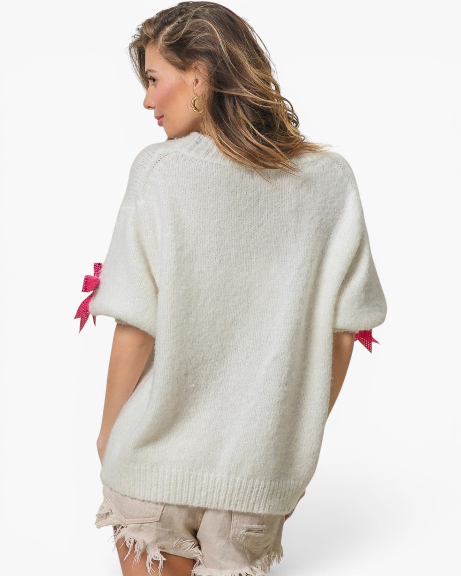 A woman shown from the back wearing a fuzzy off-white knit sweater with pink ribbon bows at the sleeve cuffs, paired with distressed shorts, against a plain white studio background.