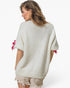 A woman shown from the back wearing a fuzzy off-white knit sweater with pink ribbon bows at the sleeve cuffs, paired with distressed shorts, against a plain white studio background.