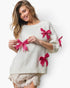 A woman wearing a cream-colored knit sweater decorated with bright pink glitter bow appliqués, paired with frayed beige shorts, against a plain white background.