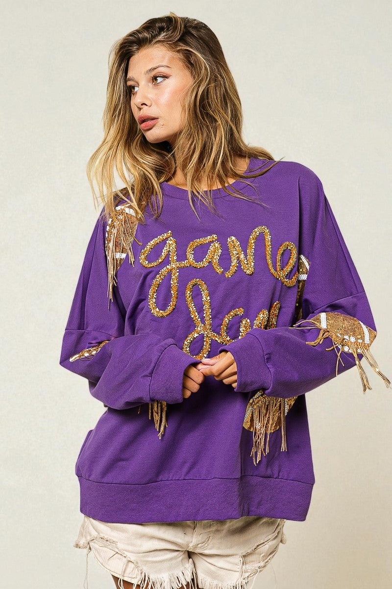 Woman wearing a purple sweatshirt with gold text and fringe details on a beige background