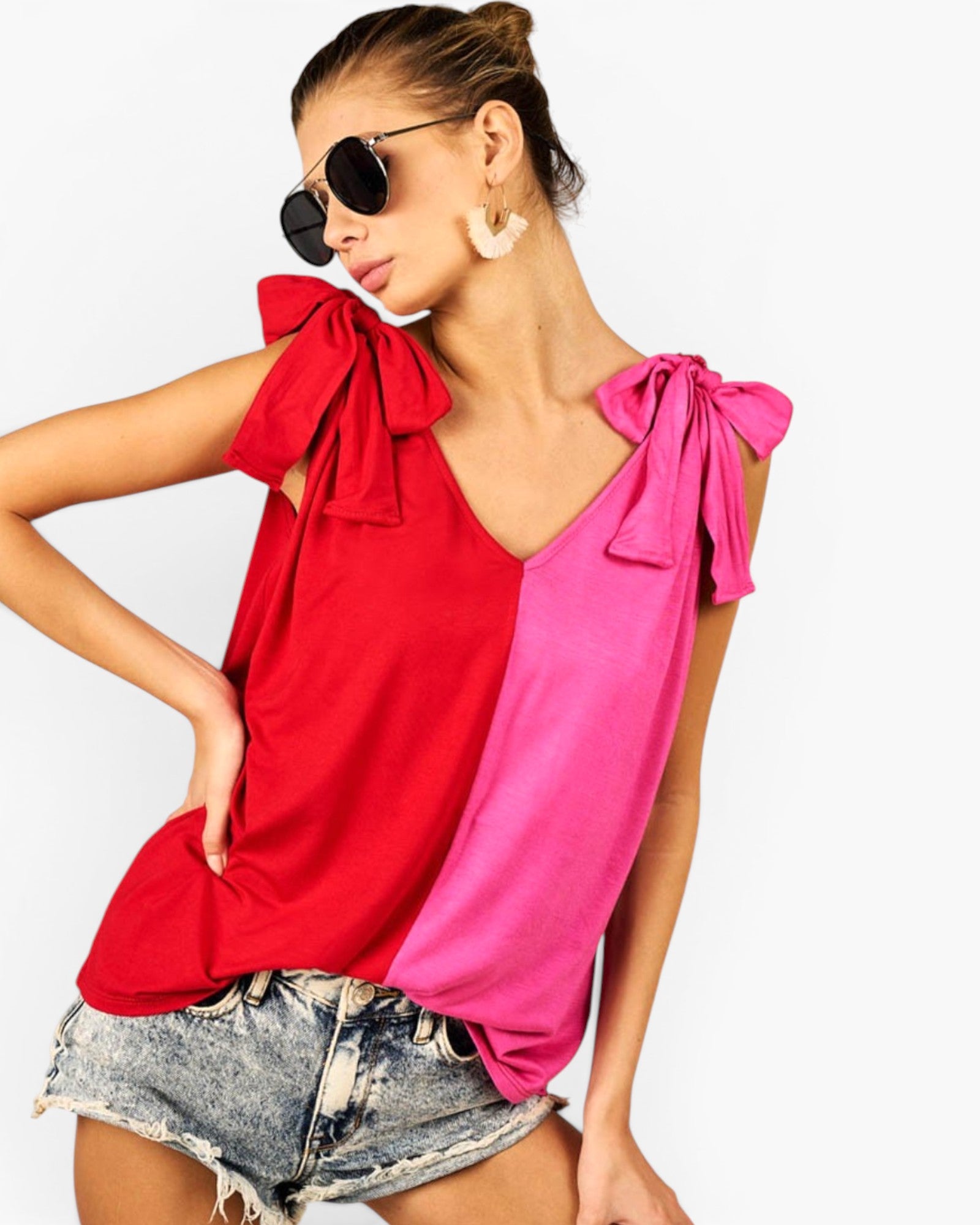 A woman modeling a sleeveless V-neck top with a split color-block design in bright red and hot pink, featuring large bow details on the shoulders, paired with denim shorts and sunglasses against a plain white background.