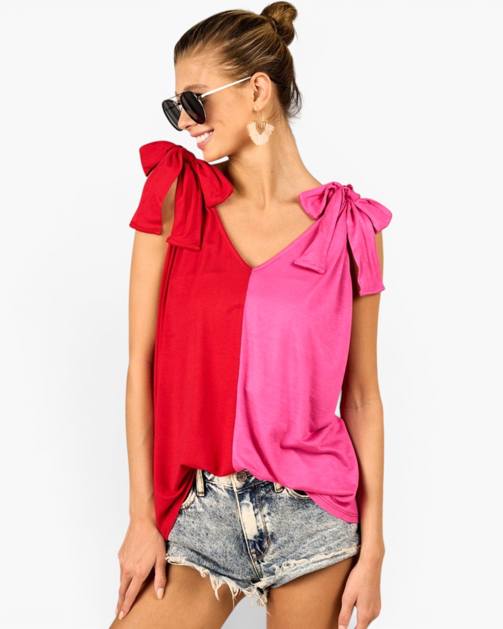 A woman modeling a split-color top with a vibrant red left side and a bright pink right side, featuring shoulder tie details and denim shorts against a plain white background.