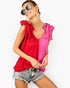 A woman wearing a sleeveless split-color top with a red left side and a bright pink right side, featuring bow details on the shoulders, paired with denim shorts against a plain white background.