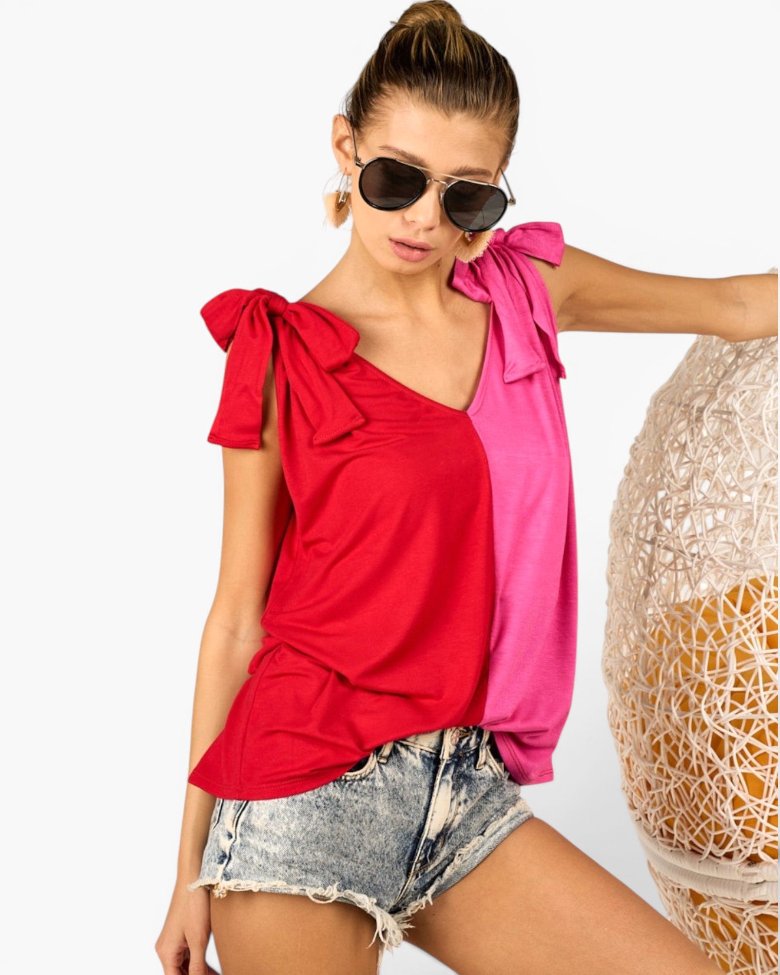 A woman wearing a split-color top with a bright red left side and a hot pink right side, styled with denim shorts and sunglasses, posed next to a woven chair against a white background.