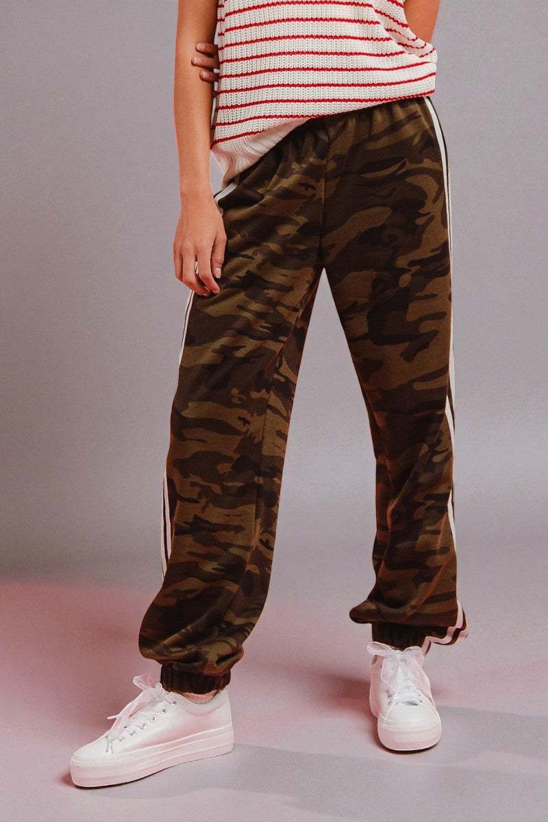 Person wearing camouflage pants with white stripes and white sneakers on a plain background