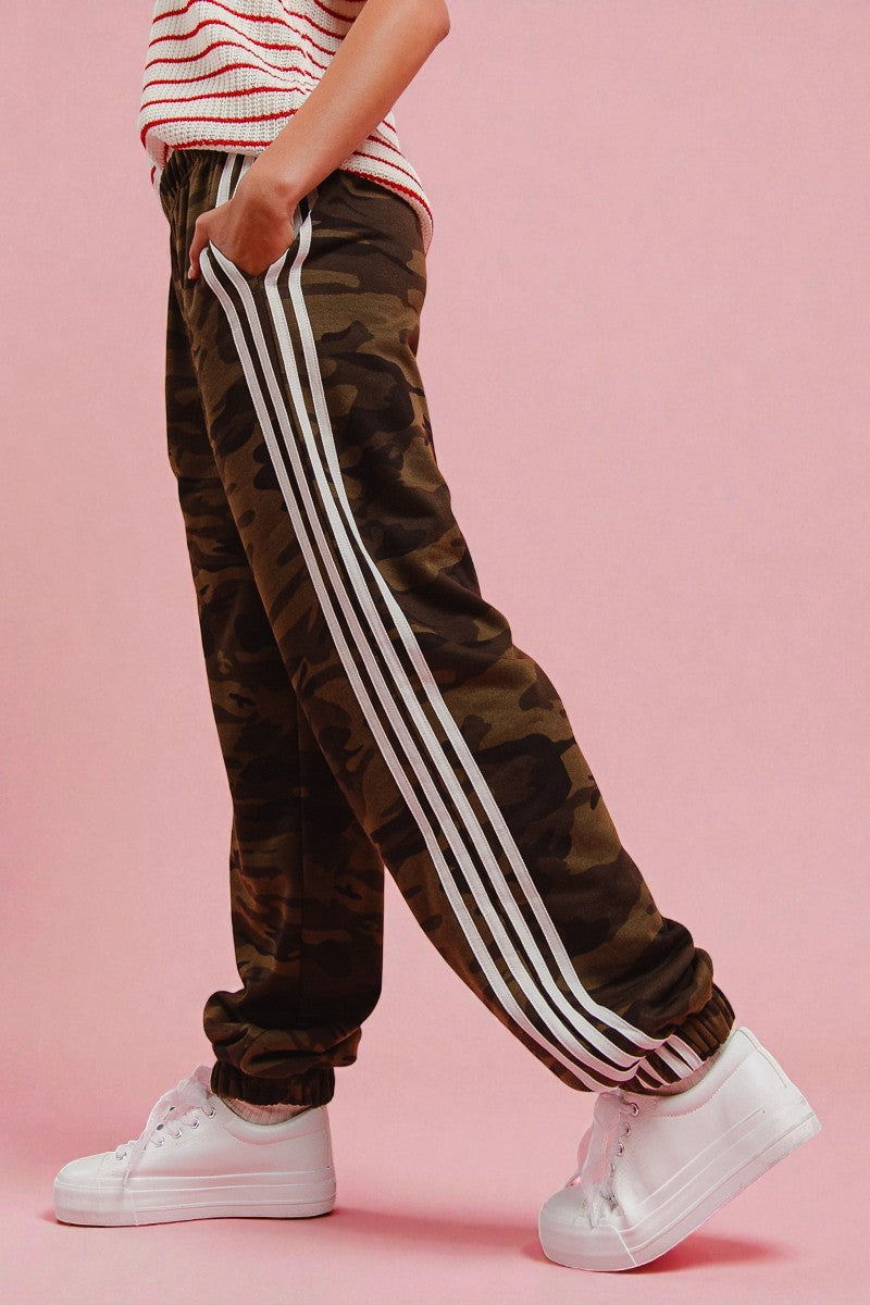 Camouflage jogger pants with white stripes worn by a person on a pink background