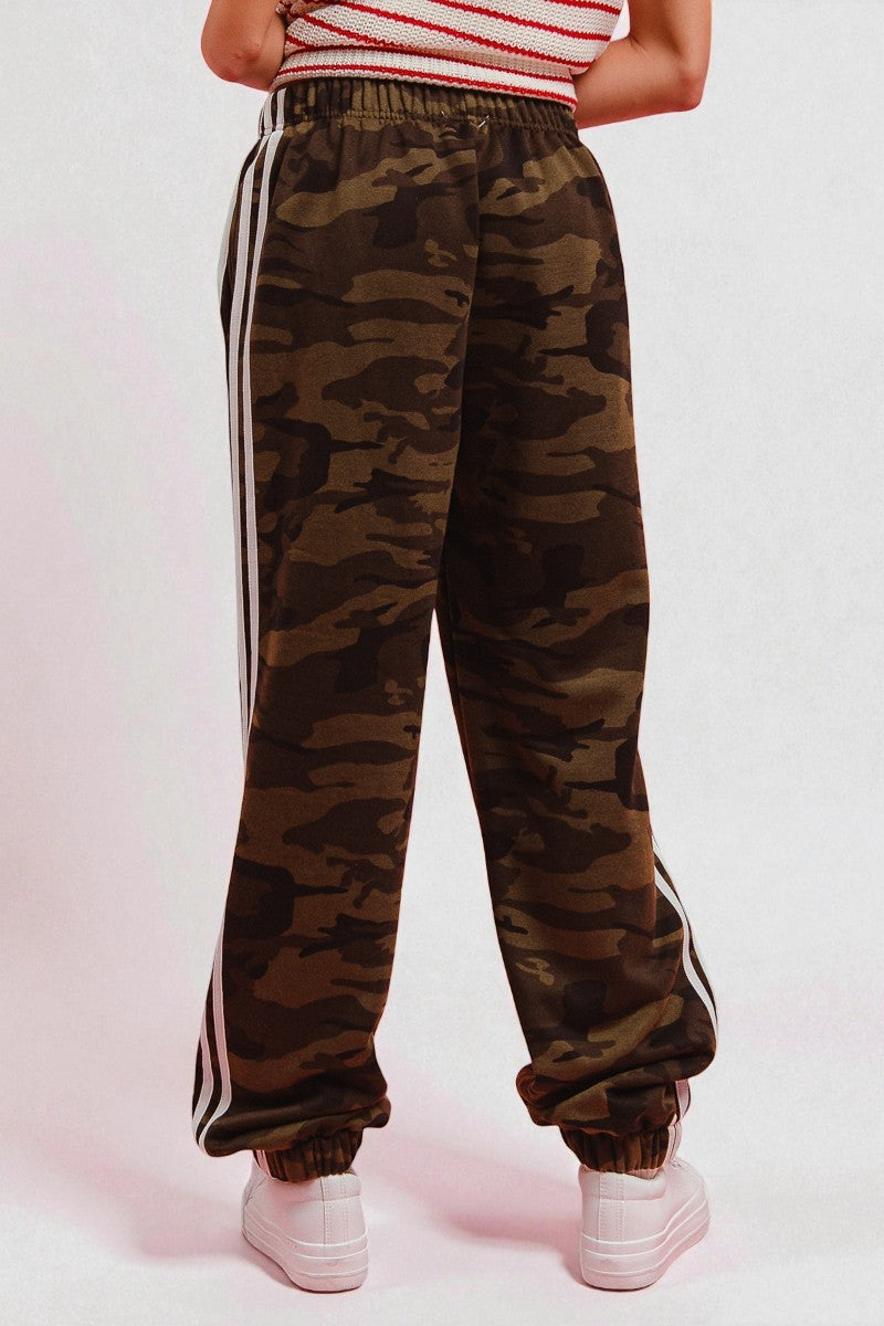 Camouflage pants with white stripes worn by a person on a light pink background