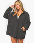 A woman modeling an oversized dark charcoal gray jacket with a button front, ribbed cuffs and hem, and contrast tan lining in the pockets, set against a plain white background.