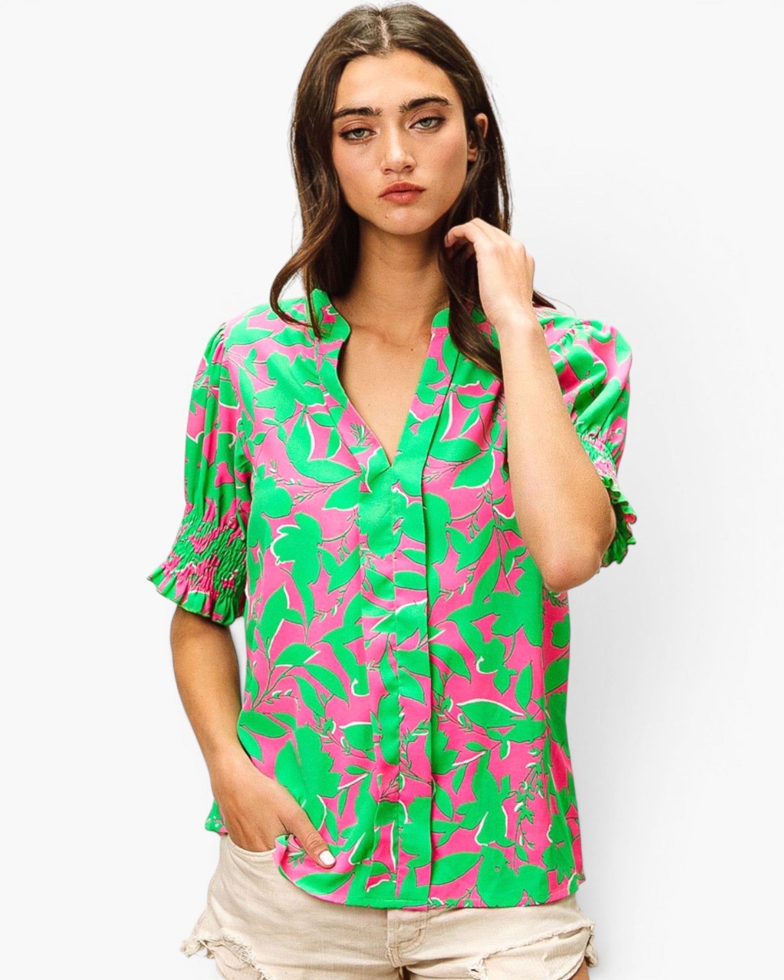 A woman modeling a short-sleeve button-up blouse with a bold green and pink tropical leaf pattern, paired with light beige shorts, against a plain light gray background.