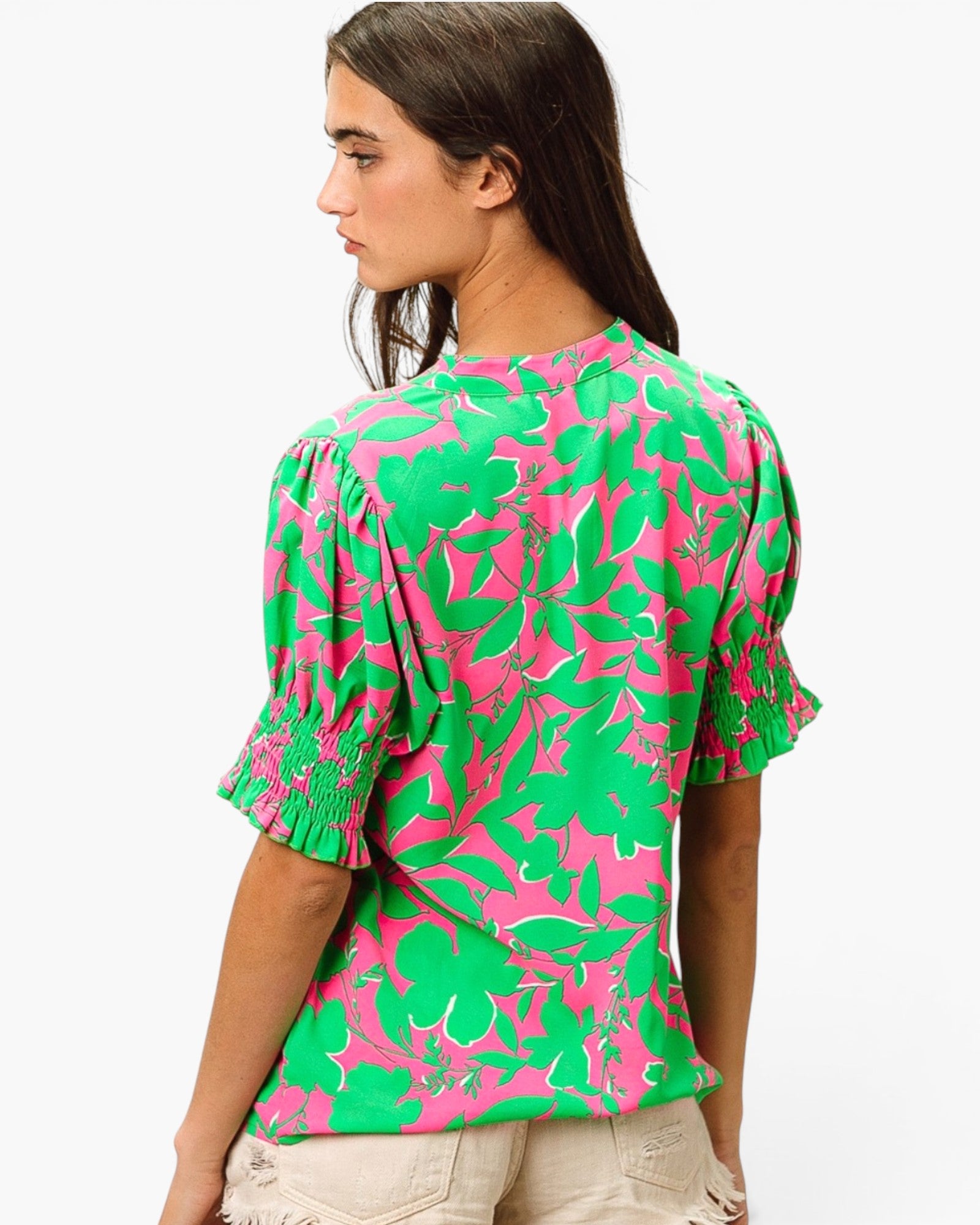 A woman shown from the back wearing a bright green and pink floral print blouse with puff sleeves and smocked cuffs, paired with light beige pants, against a plain white background.