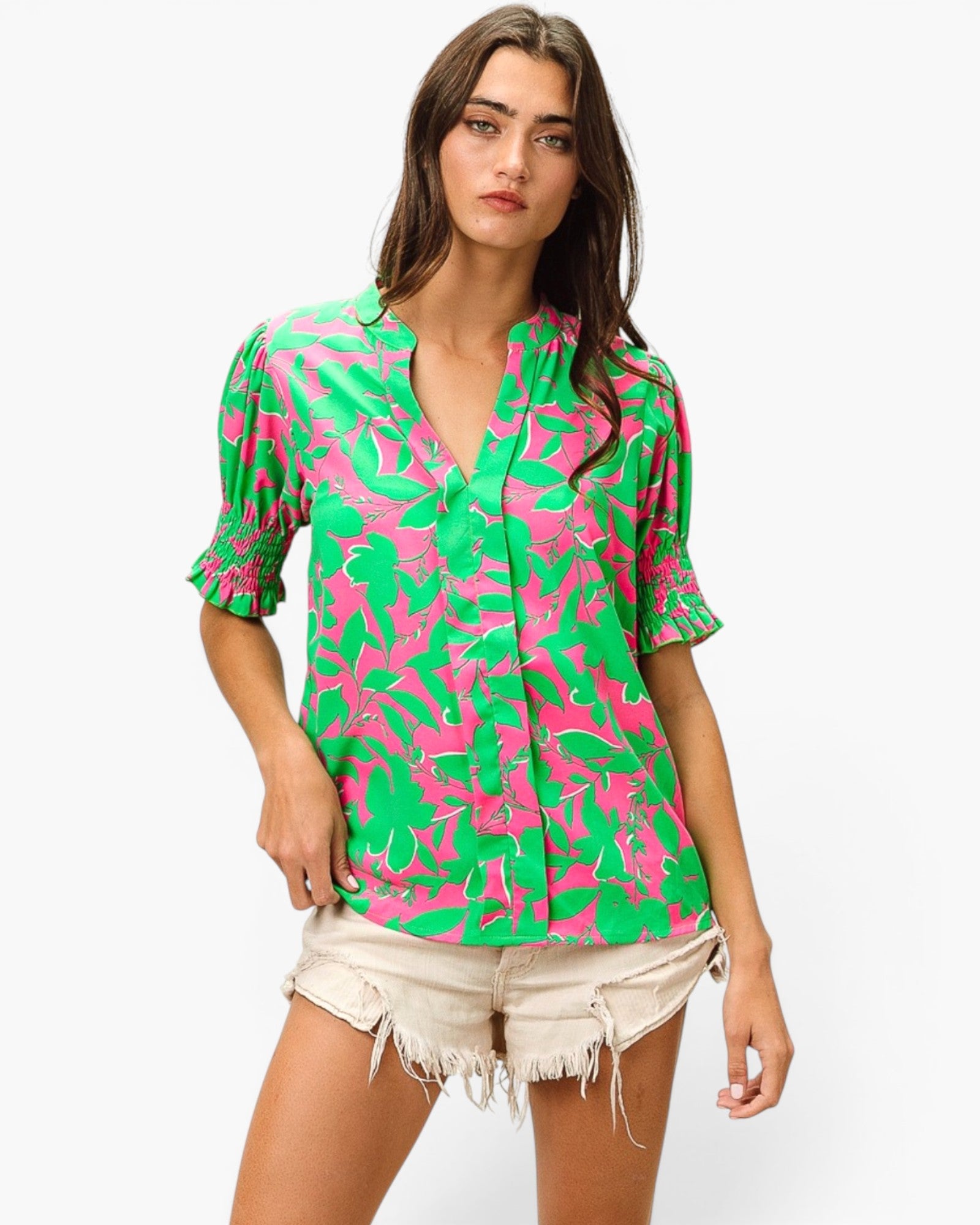 A woman modeling a bright green and pink abstract patterned short-sleeve blouse with a split neckline, paired with light beige frayed denim shorts, against a plain white background.